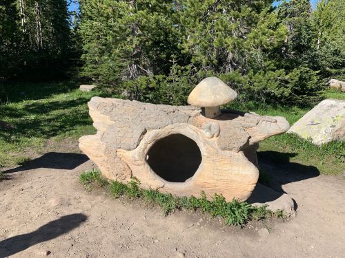 A large, carved stone resembling a log with a mushroom on top, set in a grassy area surrounded by trees.
