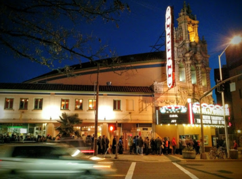 A historic theater at night, with a crowd outside and bright marquee lights illuminating the scene.