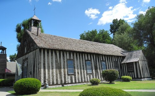 A rustic wooden church with a bell tower, featuring vertical stripes and surrounded by neatly trimmed bushes under a blue sky.