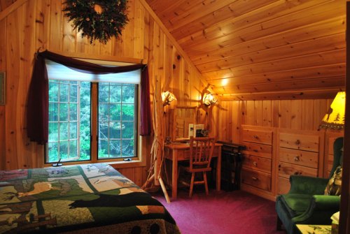 Cozy wooden bedroom with a quilted bed, desk, and large window overlooking greenery. Warm, rustic decor throughout.