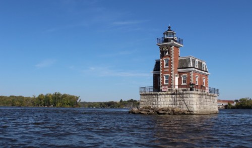 A red brick lighthouse stands on a stone base in the water, surrounded by trees under a clear blue sky.