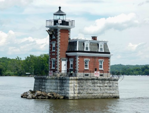 A red brick lighthouse on a stone base, surrounded by water, with a cloudy sky in the background.