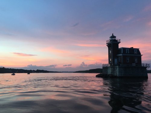 A lighthouse stands on a rocky outcrop at sunset, with colorful skies reflecting on the calm water.