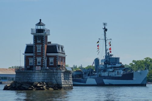 A historic lighthouse beside a large naval ship on calm waters under a clear blue sky.