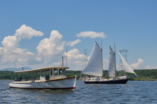 A motorboat and a sailing ship glide on calm waters under a blue sky with fluffy clouds.