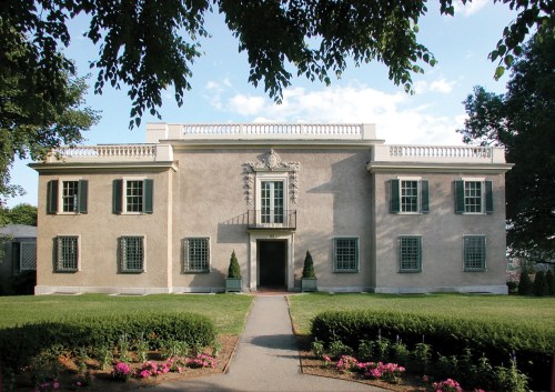 A stately, two-story building with a symmetrical facade, green shutters, and a landscaped garden in front.