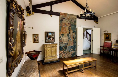 A well-lit art gallery featuring paintings, a tapestry, and wooden furniture under a high ceiling.