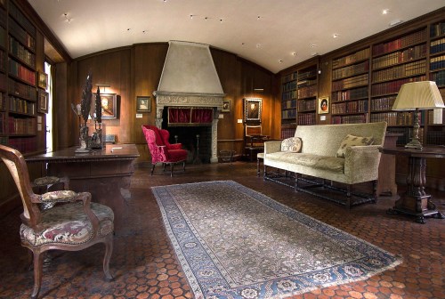 A cozy library with wooden walls, a fireplace, bookshelves, a sofa, and elegant chairs, featuring warm lighting.