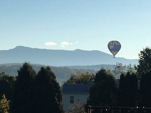 A colorful hot air balloon floats above a mountain range under a clear blue sky. Trees are visible in the foreground.