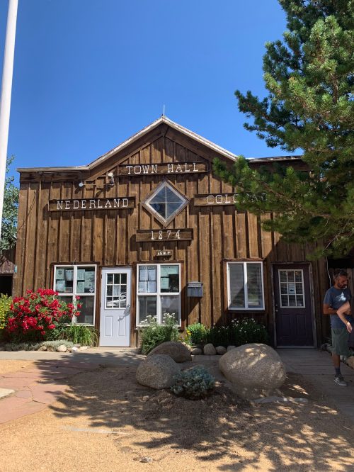 A rustic wooden building with "Town Hall" and "Nederland, Colo" signs, surrounded by greenery and flowers.