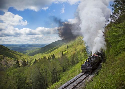 A vintage steam train chugs along a scenic mountain track, surrounded by lush greenery and a blue sky with clouds.