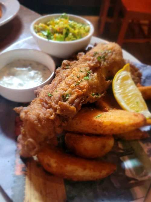 A plate of crispy fried fish with lemon, potato wedges, and a side of coleslaw and dipping sauce.