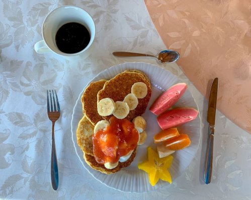 A plate of pancakes topped with bananas and strawberry sauce, accompanied by assorted fresh fruits and a cup of coffee.