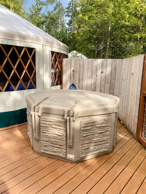A hot tub on a wooden deck next to a yurt, surrounded by trees and a wooden fence.