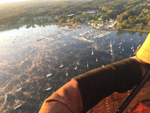 Aerial view of a marina with sailboats on a calm lake, surrounded by trees and buildings in the background.