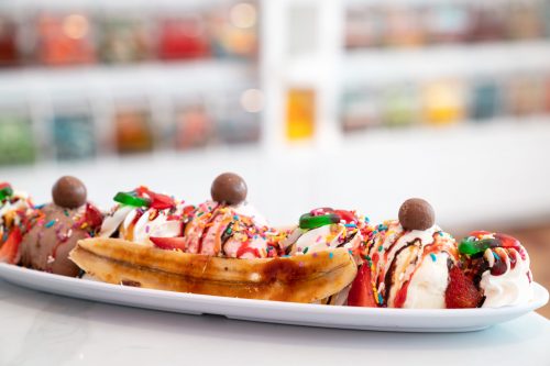 A colorful banana split topped with ice cream, whipped cream, chocolate drizzle, and sprinkles on a white plate.
