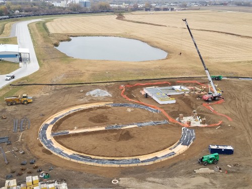 Construction site featuring a circular foundation, heavy machinery, and a crane near a pond and open fields.