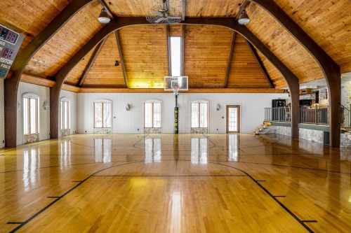 Indoor basketball court with wooden beams, polished floor, and large windows, featuring a hoop at the center.