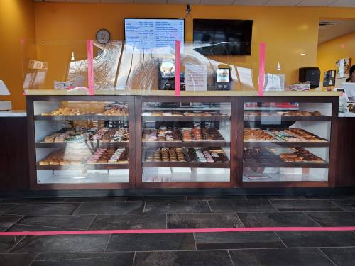 Display case filled with various donuts and pastries, with a menu board and protective glass in a bright bakery setting.