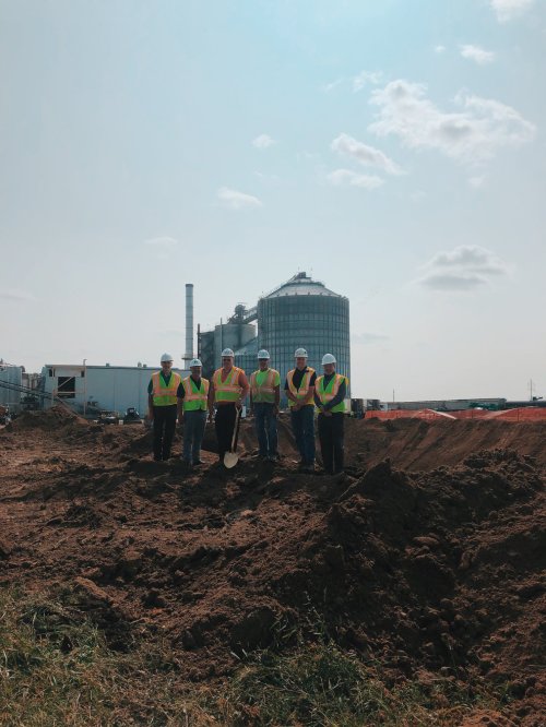 A group of six construction workers in safety vests and helmets stand on a dirt site with industrial buildings in the background.