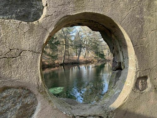 A circular stone opening frames a serene view of a river and trees in a peaceful natural setting.