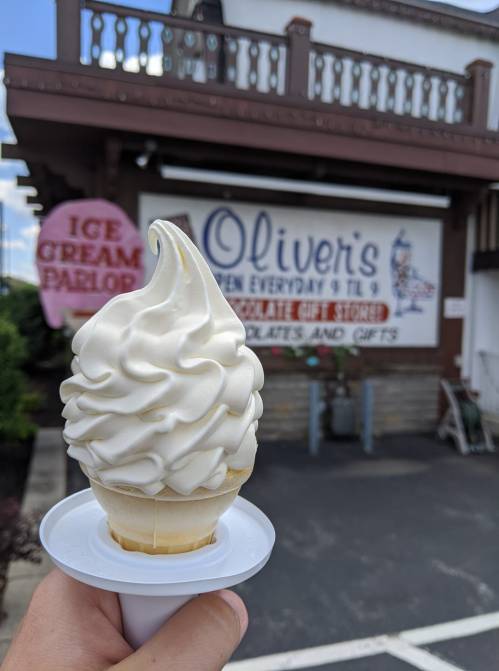 A hand holding a soft-serve ice cream cone in front of Oliver's Ice Cream Parlor sign.