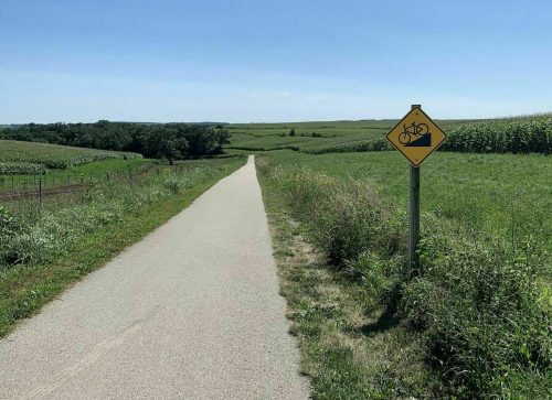 A paved path stretches into the distance, flanked by green fields and a bike warning sign under a clear blue sky.
