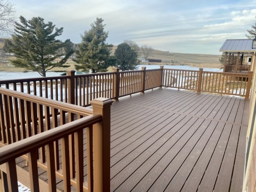 A spacious wooden deck with railings, overlooking a snowy landscape and trees under a cloudy sky.