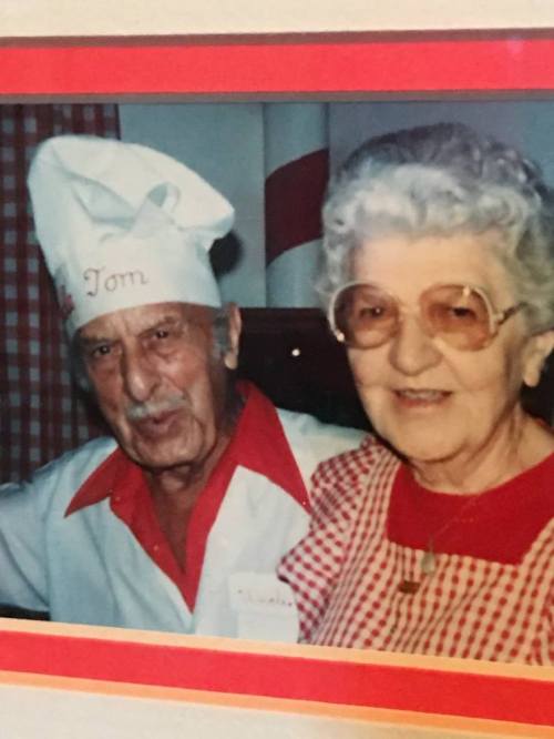 An elderly man in a chef's hat smiles next to a woman in glasses, both dressed in colorful outfits.