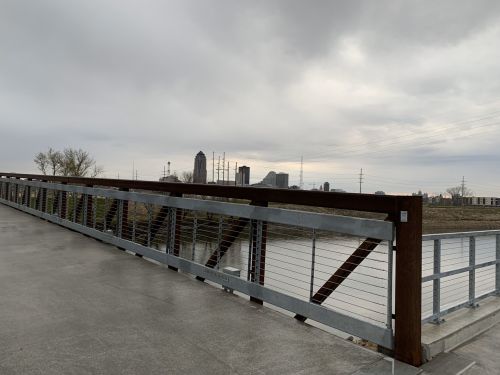 A modern bridge railing overlooks a river with a city skyline in the background under a cloudy sky.
