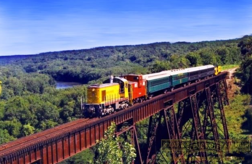 A colorful train travels across a tall bridge surrounded by lush greenery and a river in the background.