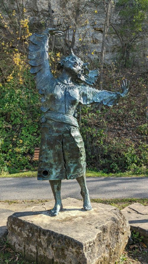 A bronze statue of a figure with wings, arms raised, standing on a rock amidst greenery and a pathway.