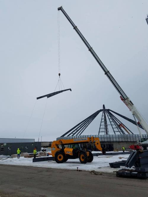 A crane lifts a steel beam at a construction site, with workers and equipment visible in a snowy landscape.