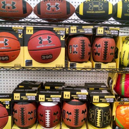 A display of various Under Armour sports balls, including footballs, basketballs, and soccer balls, on store shelves.