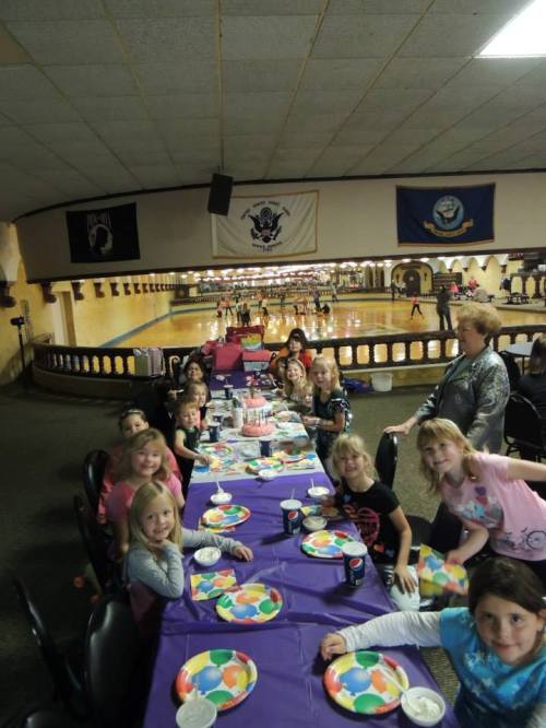 A group of children at a birthday party, sitting at a long table with colorful plates and decorations in a roller rink.