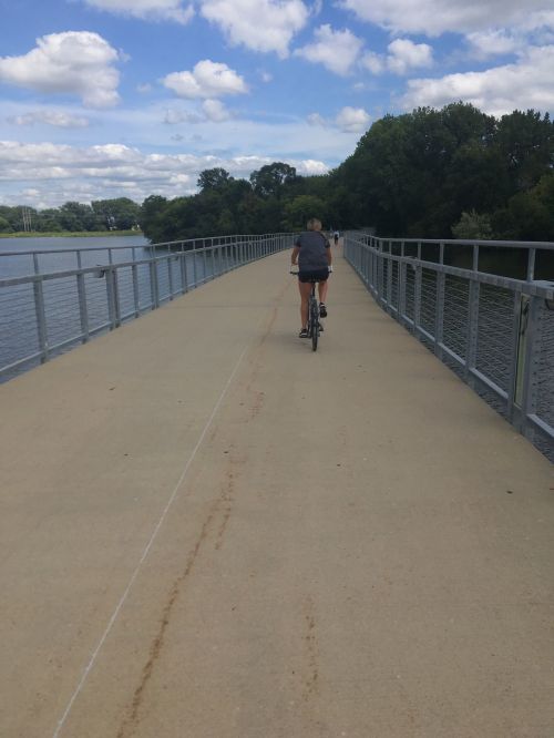 A person rides a bicycle on a wide, paved path beside a calm body of water under a blue sky with fluffy clouds.