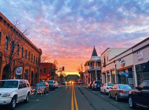 A vibrant sunset casts colorful clouds over a charming street lined with parked cars and historic buildings.