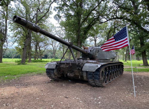 An old tank displayed in a park, with American flags on either side, surrounded by trees.