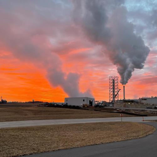 Industrial scene at sunset, featuring smokestacks emitting steam against a vibrant orange and pink sky.