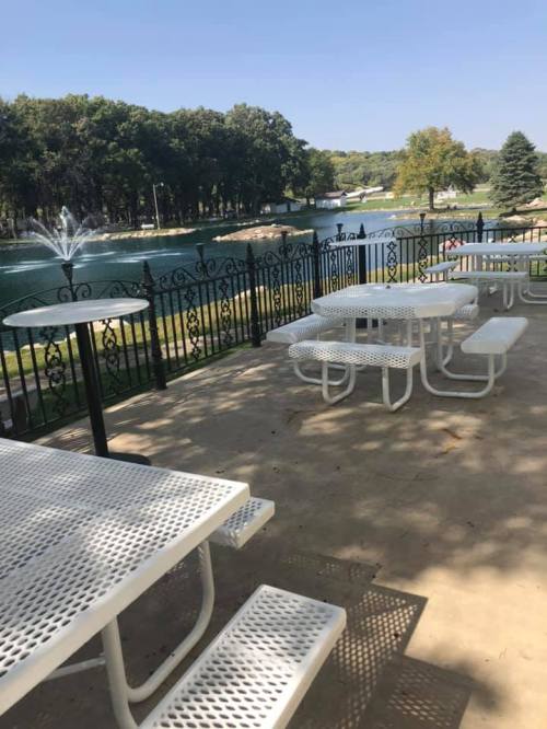 A serene outdoor area with white picnic tables overlooking a lake and fountain, surrounded by trees.