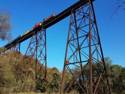 A colorful train travels across a tall, rusted trestle bridge against a clear blue sky and green trees below.