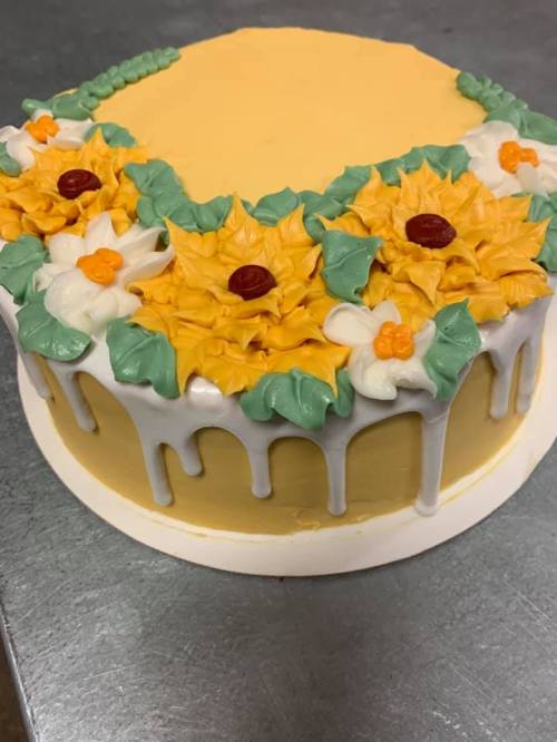 A round cake with yellow frosting, decorated with orange flowers, green leaves, and white icing drips.