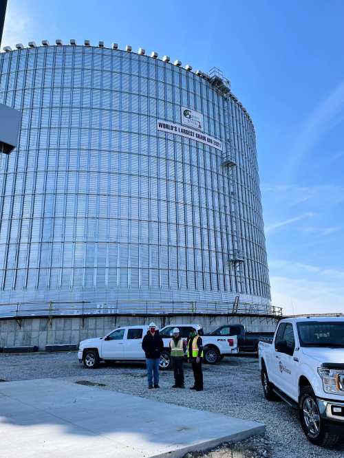 Construction site featuring a large grain storage facility with workers discussing plans and vehicles nearby.