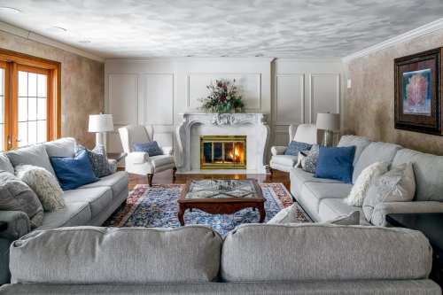 Cozy living room with a fireplace, light gray sofas, decorative pillows, and a colorful rug. Natural light from windows.