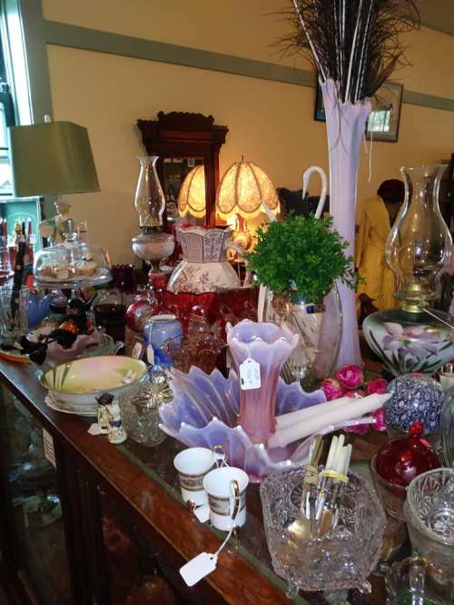 A display of various vintage glassware, lamps, and decorative items on a wooden table in a cozy shop.