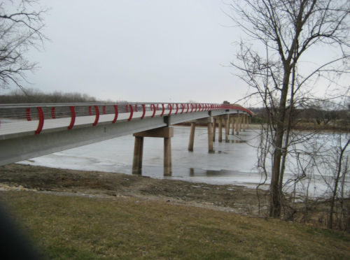 A modern bridge with red railings spans a river, surrounded by bare trees and a grassy bank.