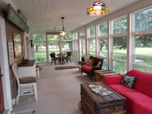 A bright sunroom with large windows, a red couch, wooden furniture, and a dining table, surrounded by greenery.