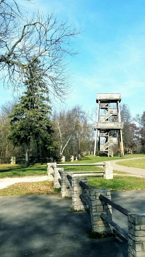 A wooden observation tower stands in a park, surrounded by trees and a clear blue sky. Pathways lead through the area.