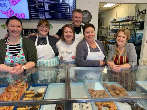 A group of five smiling bakers stands behind a display of assorted pastries in a bakery.