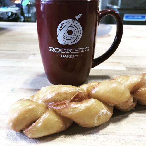 A braided pastry next to a maroon mug with "ROCKETS BAKERY" printed on it, set on a wooden table.